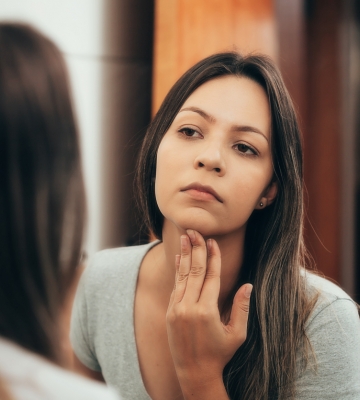 A woman in her thirties, long brown hair looking in the mirror to do a thyroid self-exam.